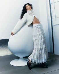 Olivia Dean poses in a cropped off-white cardigan and tiered ruffled maxi skirt while leaning on a white sculptural pod chair.