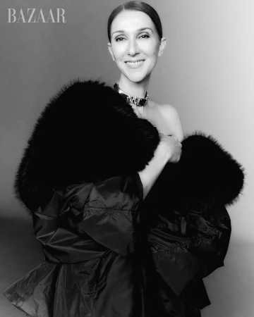 Celine Dion smiling in a dramatic black fur-trimmed wrap coat and metallic choker necklace against a neutral studio backdrop.