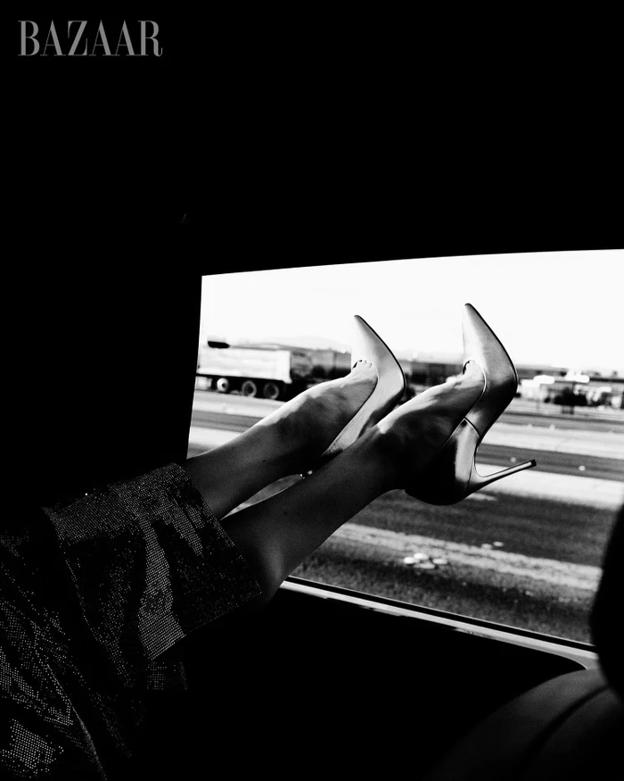A person's legs wearing pointed-toe stiletto pumps extended out of an open car window along a highway in black and white.
