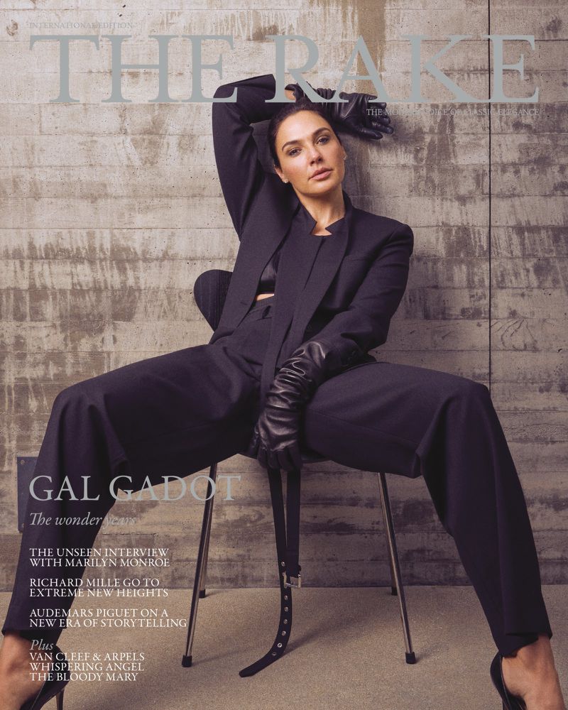 Gal Gadot seated on a modern chair against a concrete wall wearing a tailored black suit and black leather opera gloves.