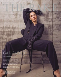 Gal Gadot wearing an oversized black suit and long leather gloves seated on a chair against a concrete wall for The Rake magazine cover.