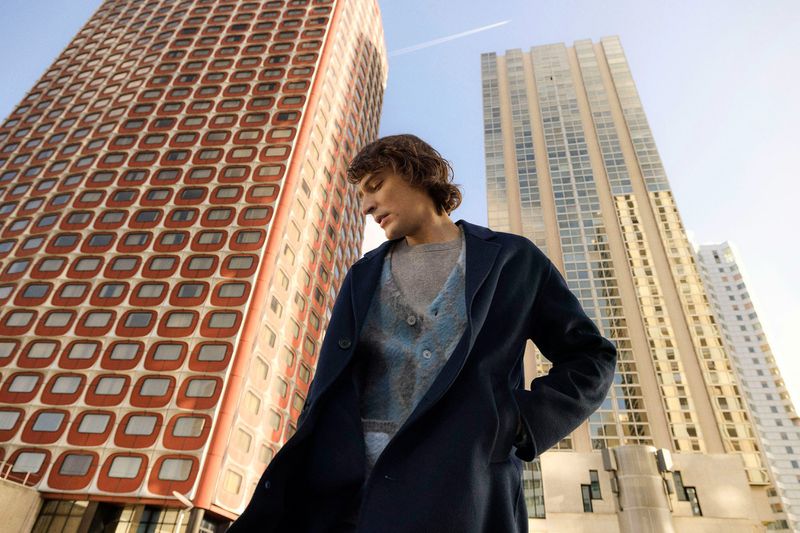 A man with wavy hair wearing a navy wool coat and blue patterned cardigan, standing outside against tall, modern urban skyscrapers.