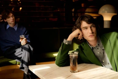 A man in a green blazer leans on a marble table with an iced drink while another man sits in a lounge background.