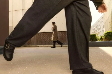 Low-angle view framed between walking legs in charcoal trousers showing a man walking in a long beige coat in the background.