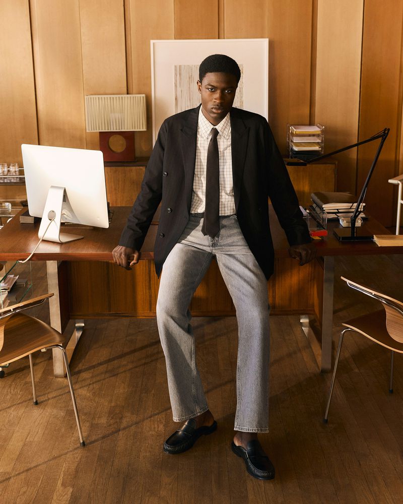 A young Black man wearing a black blazer, white grid shirt, tie, and light grey jeans, sitting on a desk in a wooden office.