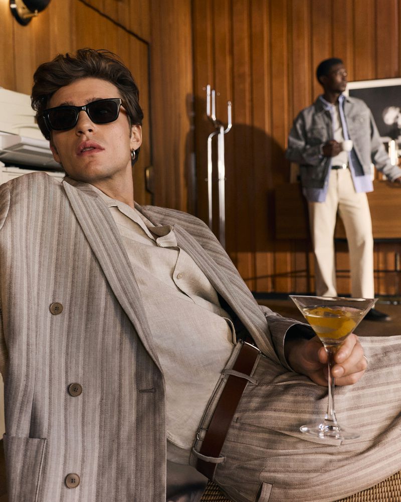 Man in dark sunglasses and a beige striped suit holding a cocktail glass, with another man standing in a wood-paneled room behind him.