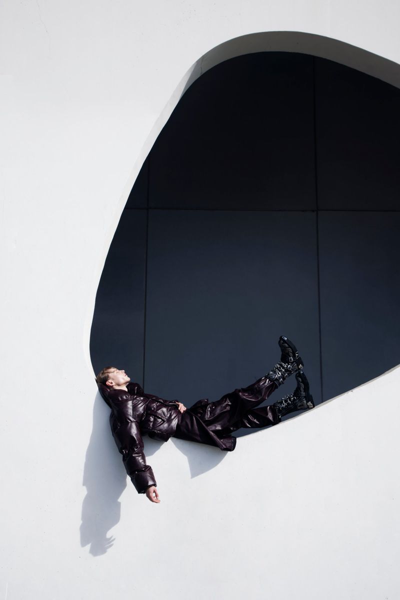Man wearing a glossy brown puffer jacket and matching trousers reclining against the edge of a curved, modern white architectural cutout.