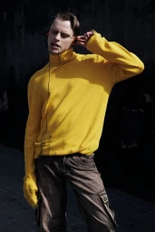 Man in a distressed bright yellow turtleneck sweater and cargo pants holding wired headphones in direct sunlight.