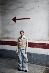 Fashion model in a beige zip jacket and oversized distressed denim cargo pants standing beneath a painted red arrow on a wall.