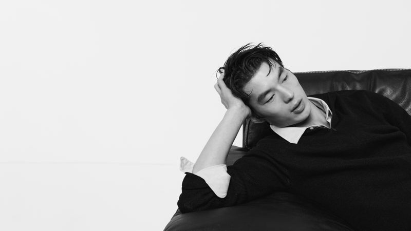Young man with dark hair in a white shirt and black sweater resting on a dark leather sofa in a minimal studio setting.