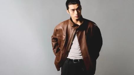 Model Xiangyu Li posing in a brown leather jacket over a grey henley shirt against a neutral grey studio backdrop.