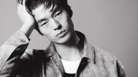 Black and white portrait of model Xiangyu Li posing with a hand on his head wearing a collared overshirt against a grey background.