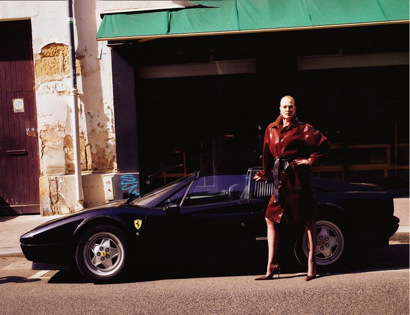 Model Karen Mulder in a glossy brown coat standing next to a dark sports car parked on a sunny city street.
