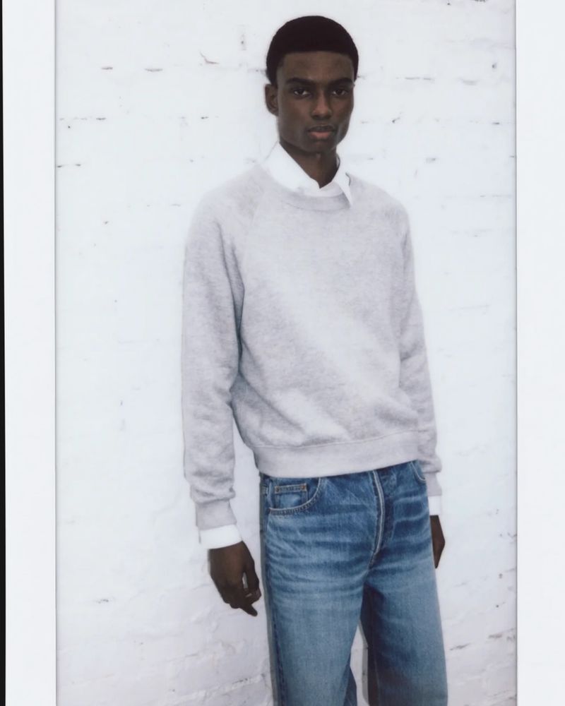 A young Black man wearing a light grey crewneck sweatshirt over a white button-down shirt and blue jeans against a white brick wall.