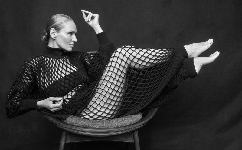 Black and white portrait of an adult woman in a chair wearing a net-like dress in a photo studio against a dark backdrop.