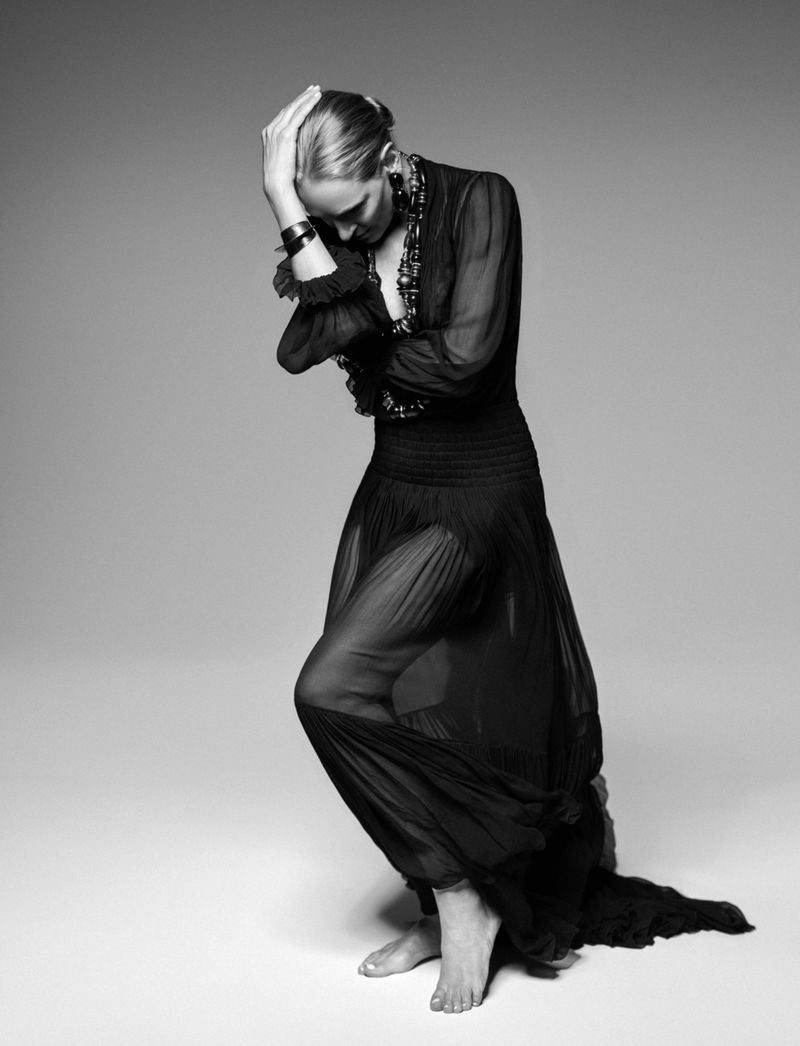 Black and white portrait of Uma Thurman posing barefoot in a sheer pleated black gown against a studio seamless background.
