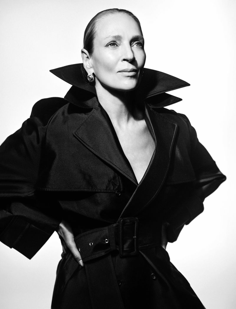 Black and white portrait of Uma Thurman posing in a belted black trench coat with an exaggerated collar against a white background.