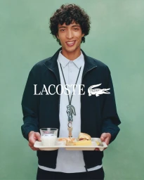 A smiling man with curly hair holds a cafeteria food tray with a glass of milk and sandwich against a green background.
