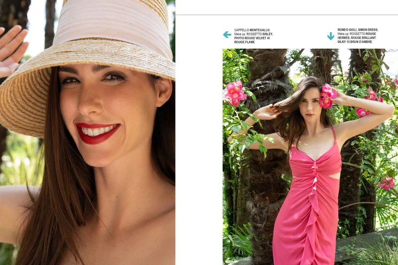 Model in a pink dress wearing a straw hat poses outdoors among lush greenery and pink flowers in bright, natural lighting.