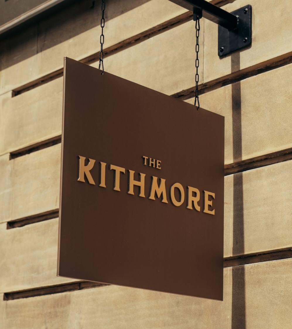 Square brown hanging sign with raised gold lettering reading THE KITHMORE suspended by black chains on a stone facade.