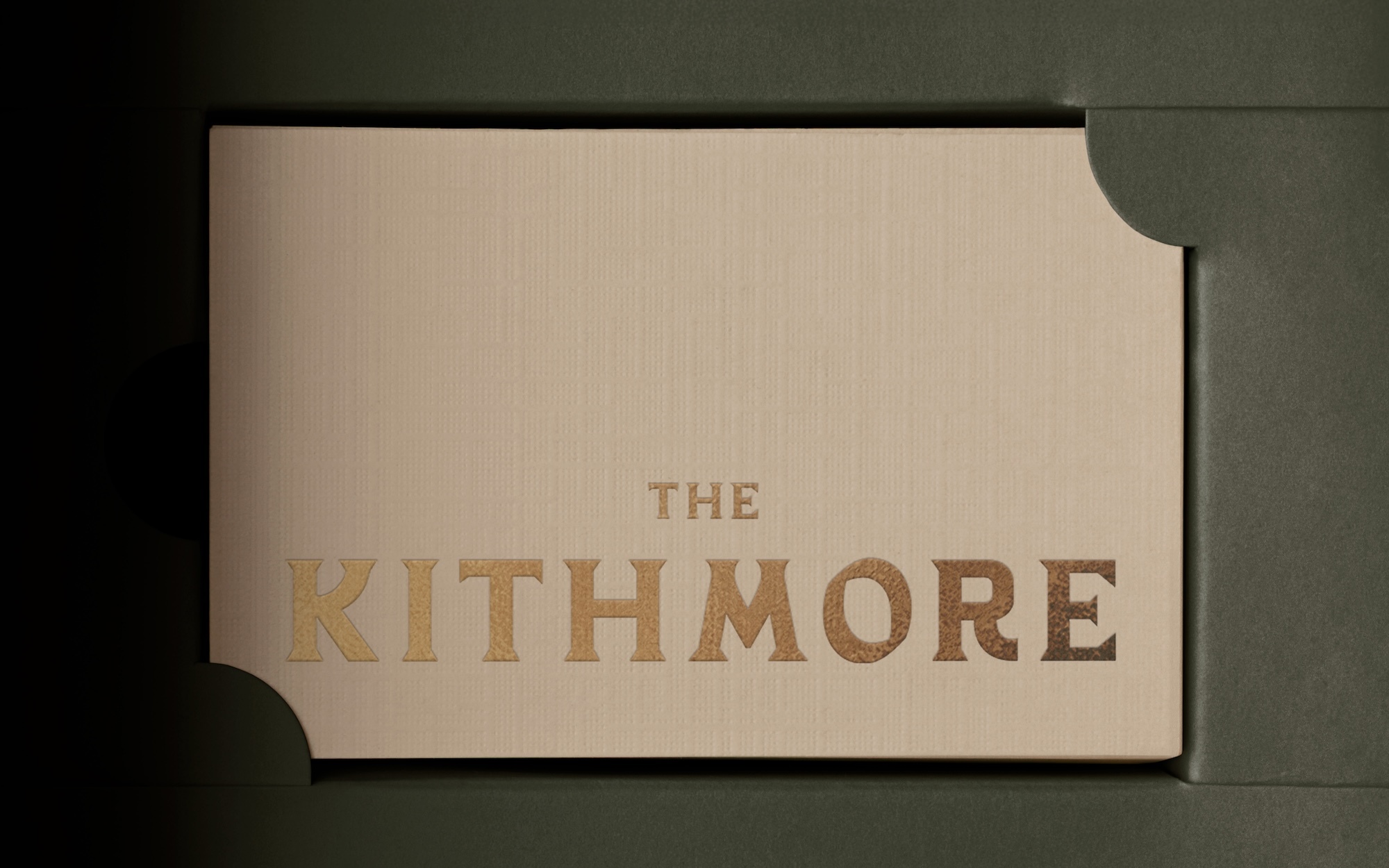 The Kithmore gold typography on textured beige card stock slotted into a dark olive green packaging sleeve.