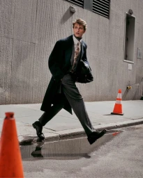 Model Vasko Luyckx strides along a city sidewalk wearing a dark tailored overcoat, grey trousers, and a patterned tie.