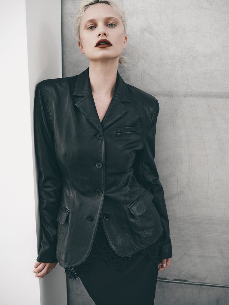 Model wearing a structured black leather blazer with dark lipstick leaning against a grey concrete wall.
