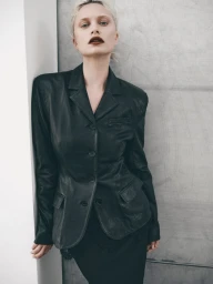 Model wearing a structured black leather blazer with dark lipstick leaning against a grey concrete wall.