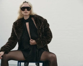 Blonde woman in dark sunglasses and a leopard-print fur coat seated on a chair against a white background.
