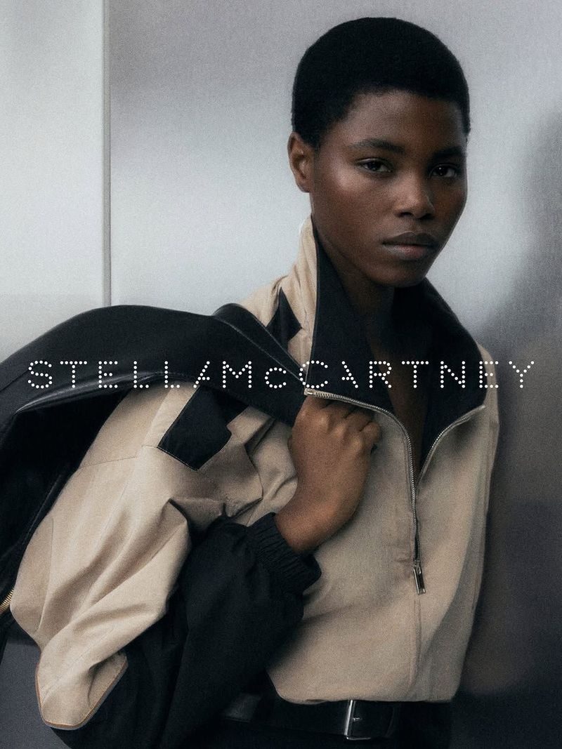 A person with short hair wears a beige and black high-neck zip jacket, looking directly at the camera against a metallic background.