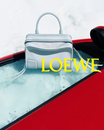 Light blue Loewe leather handbag resting on the wet windshield of a red car surrounded by snow with yellow logo typography.