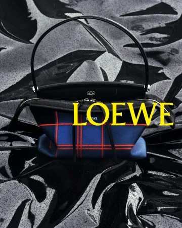 Loewe plaid handbag with curved black top handle resting on a reflective black liquid surface with yellow logo overlay.