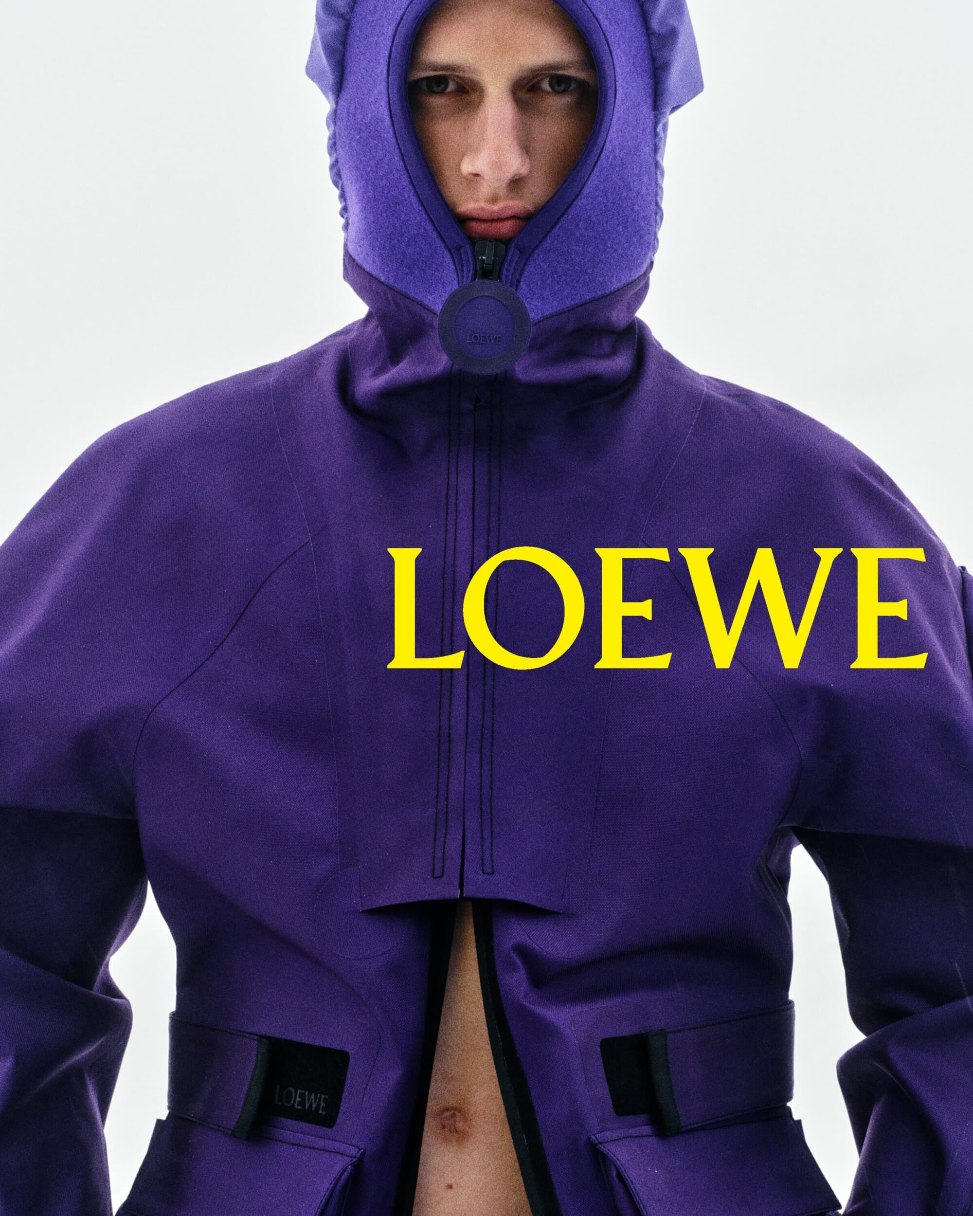 Model wearing a structured purple hooded technical jacket with yellow Loewe typography across the chest against a pale sky.