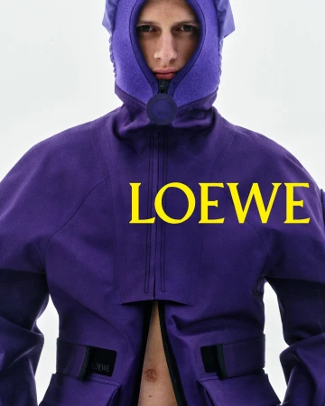 Model wearing a structured purple hooded technical jacket with yellow Loewe typography across the chest against a pale sky.