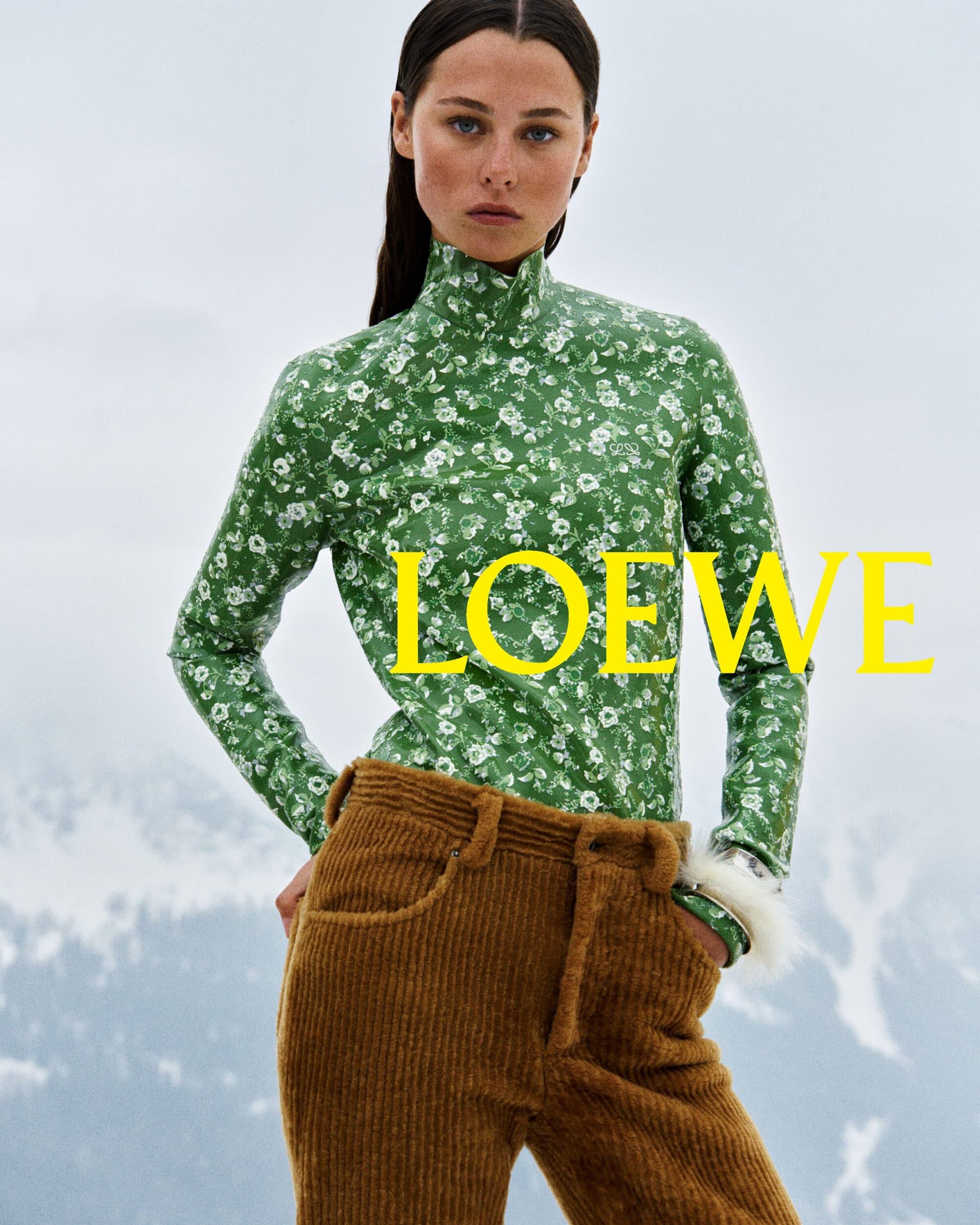 Model wearing a green floral high-neck top and brown corduroy trousers against a snowy mountain landscape.