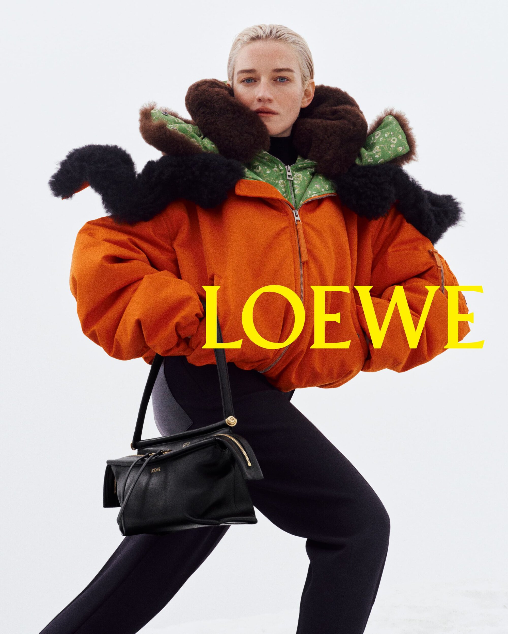 Model wearing an oversized orange bomber jacket with a fur collar and black leggings holding a black Loewe leather handbag on snow.