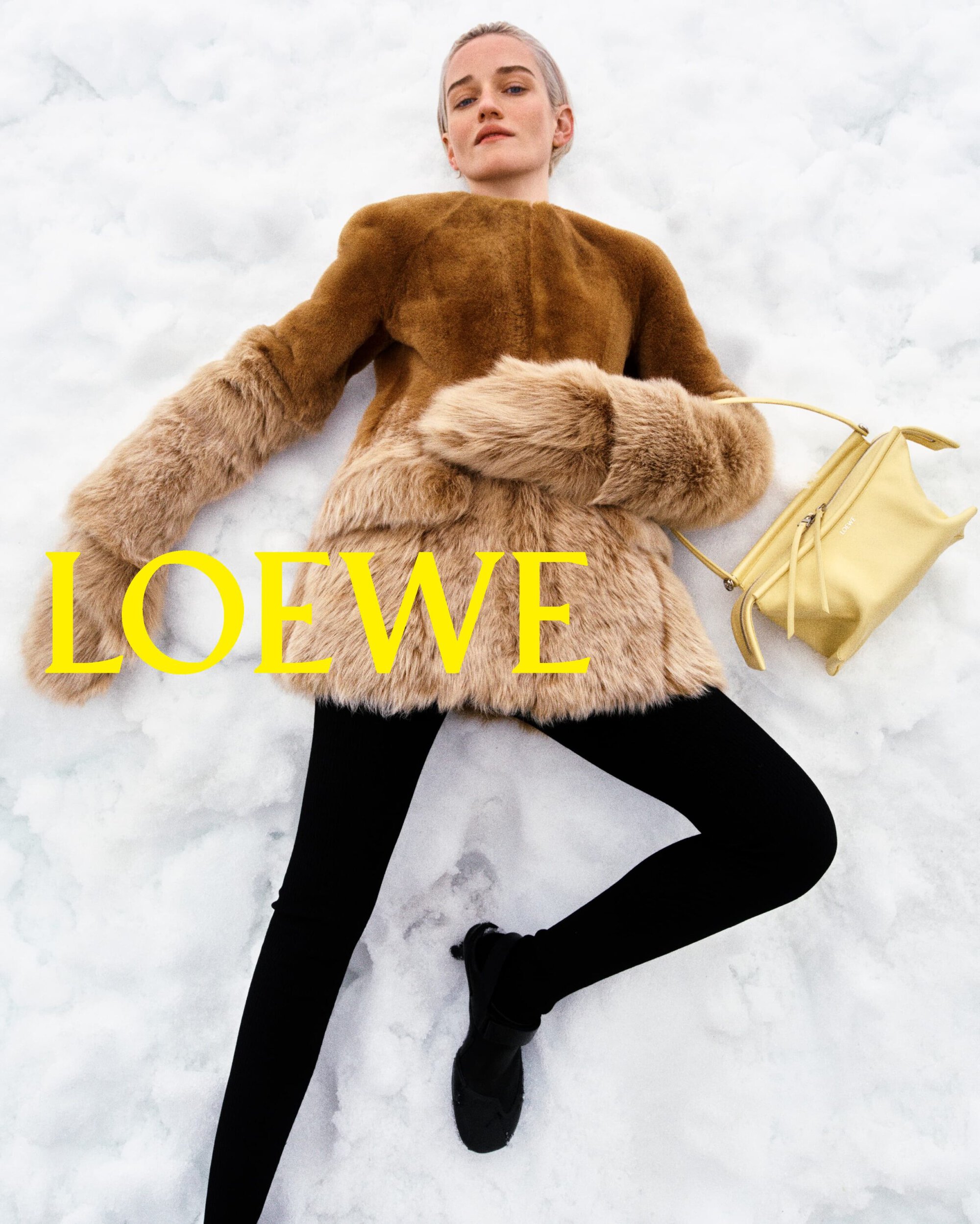 Model lying on snowy ground wearing a brown plush shearling coat, black leggings, and holding a pale yellow Loewe handbag.