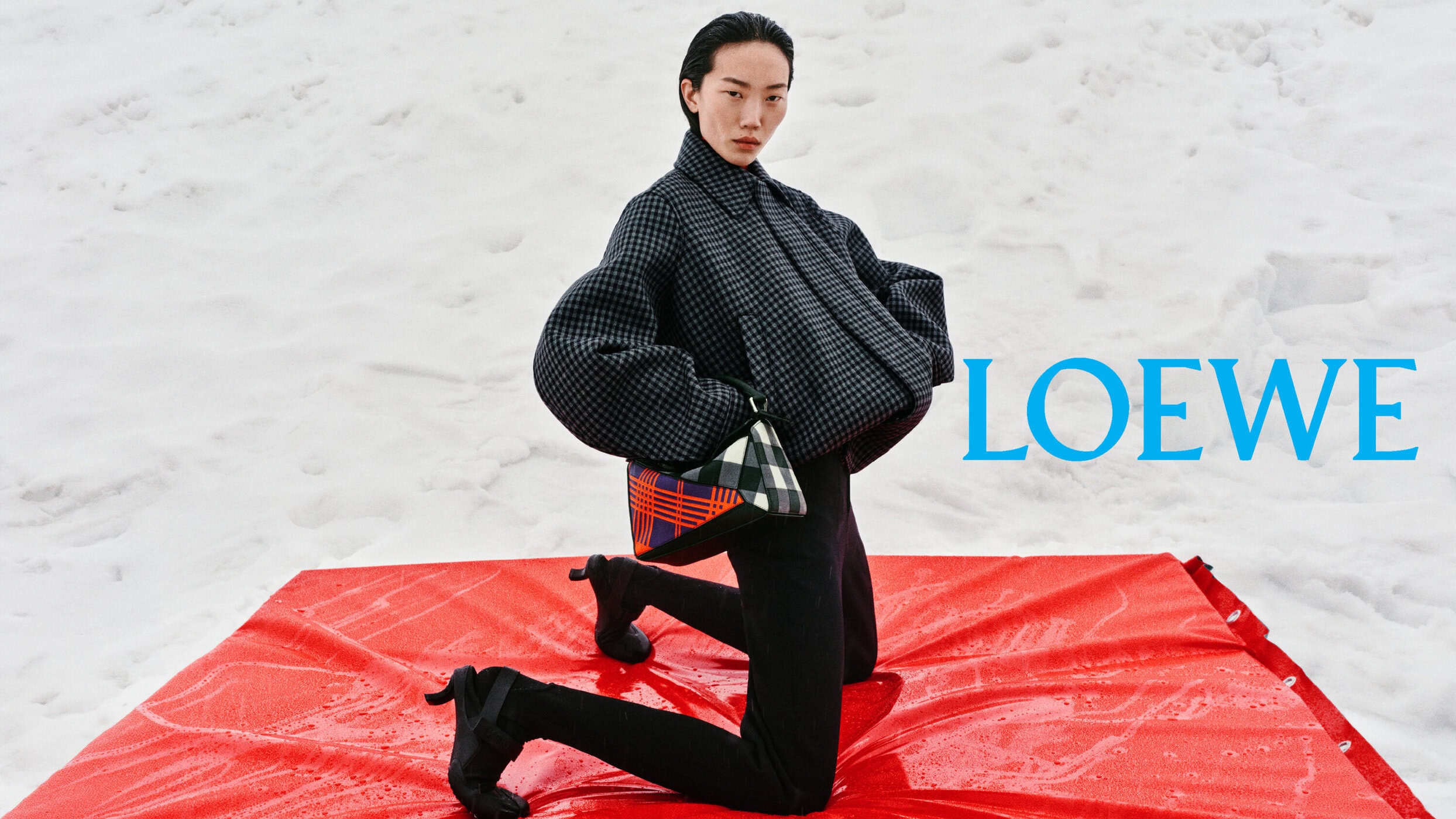 Model in an oversized grey checked jacket kneeling on a bright red tarp in the snow with a Loewe handbag.