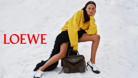 Model in a bright yellow sweater and textured black skirt posing in the snow with a dark suede Loewe handbag.