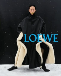 Fashion model in an oversized black coat with cream shearling lining standing on snow with a vibrant blue Loewe logo across the frame.