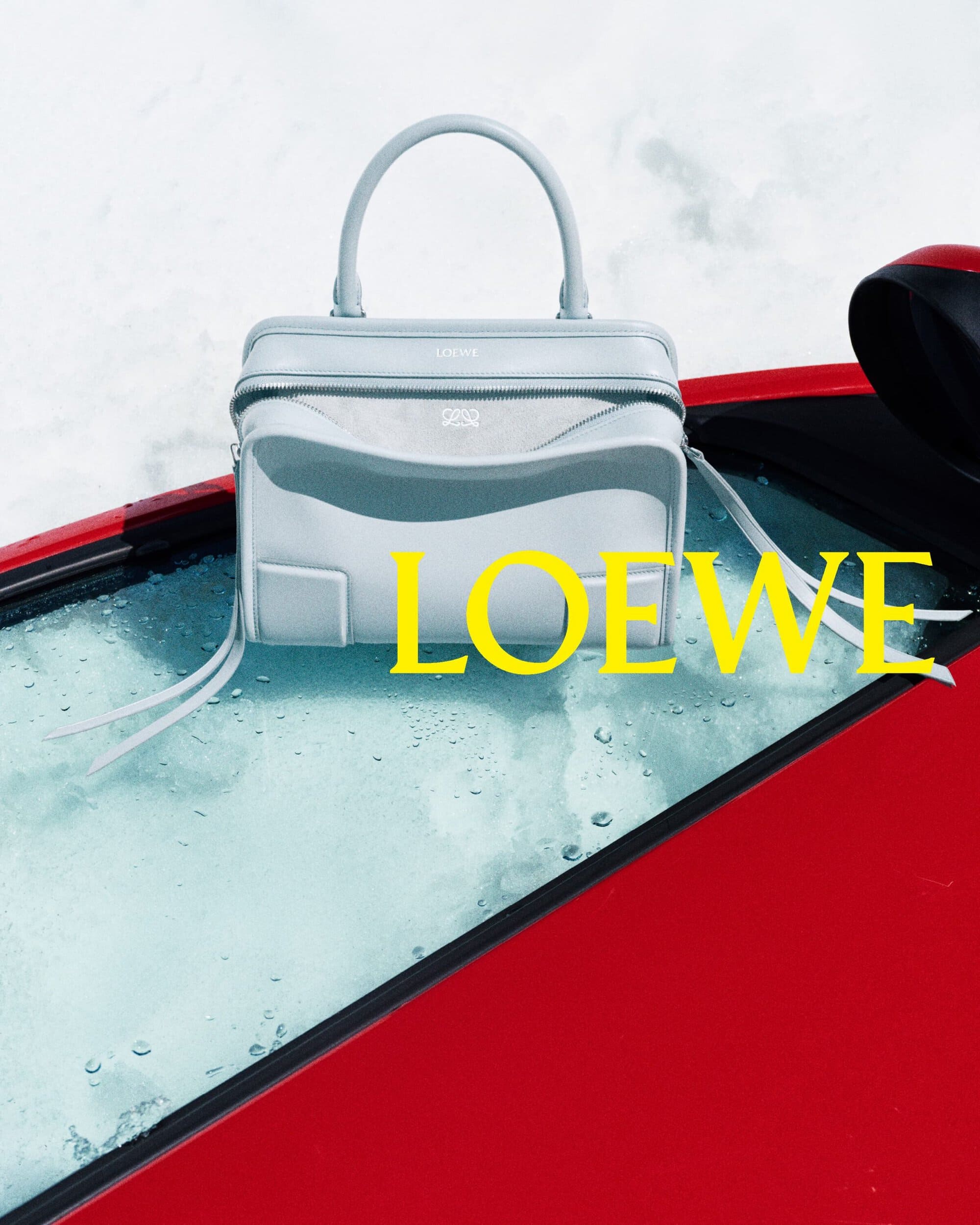 A pale blue Loewe leather handbag rests on the wet glass roof of a red car in a snowy alpine setting with yellow brand typography.