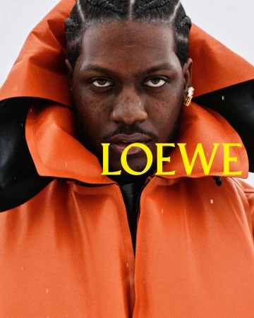 Close-up portrait of Lil Yachty wearing a bright orange hooded jacket with yellow Loewe typography across the collar.