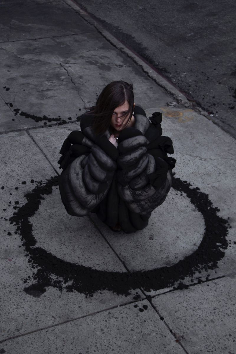 Young woman with dark hair kneels in a dirt circle on gray pavement. She wears a gray and black coat in an outdoor urban environment.