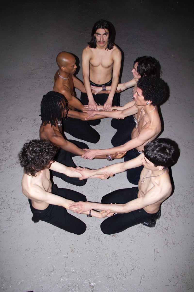 Overhead shot of a group of shirtless young adult men in dark pants, forming a circle, holding hands in a dark gray indoor setting.