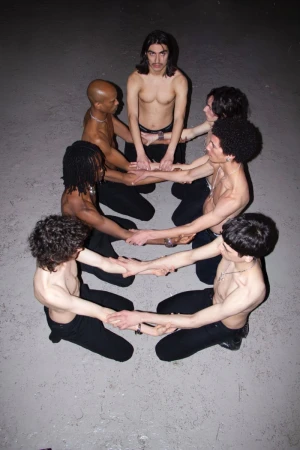 Overhead shot of a group of shirtless young adult men in dark pants, forming a circle, holding hands in a dark gray indoor setting.