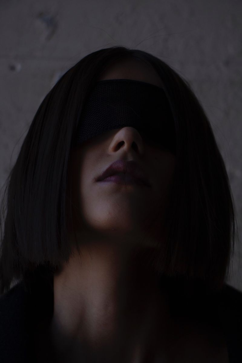 Portrait of a person with short dark hair wearing a black blindfold. The photo is close-up and shows the head and shoulders.