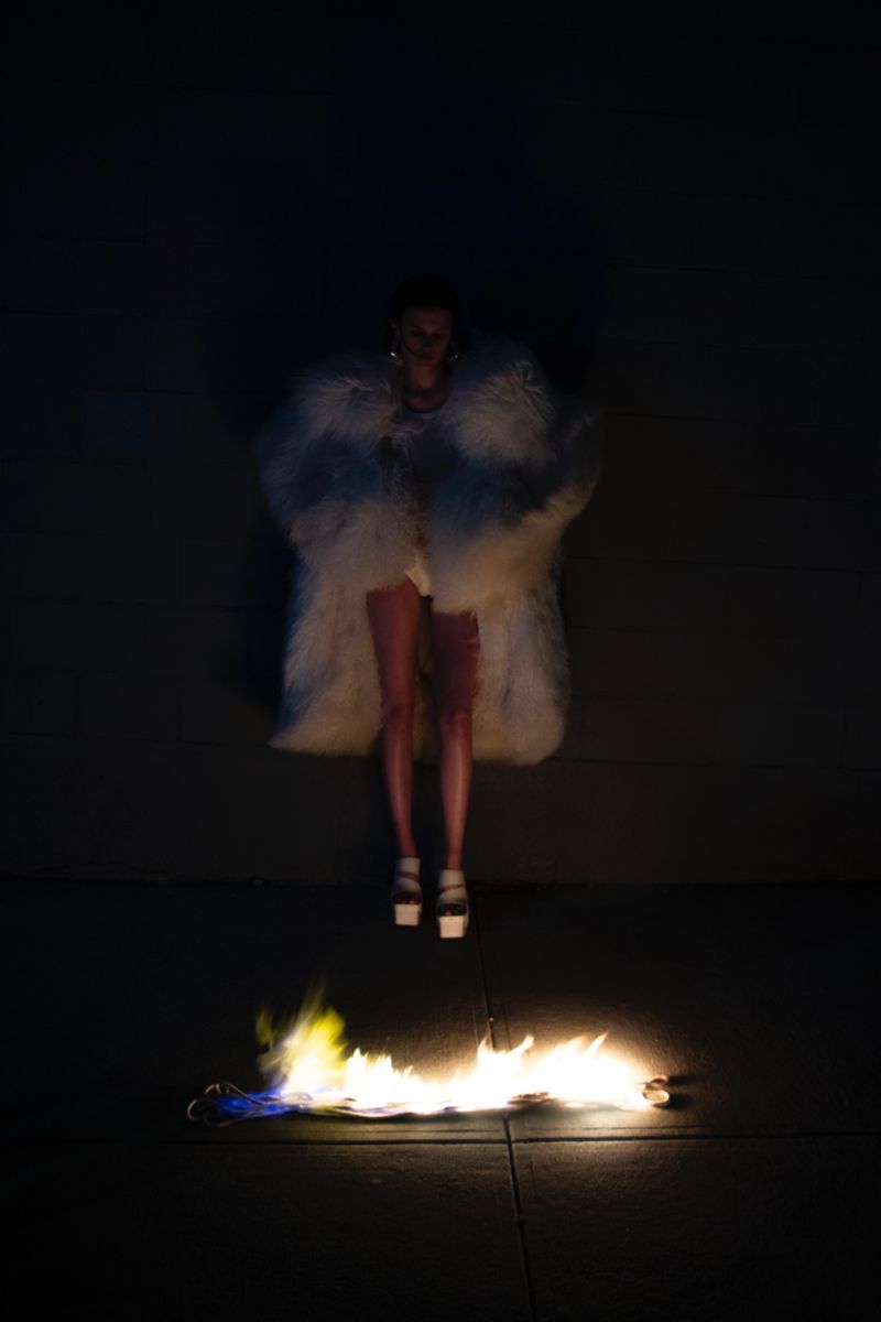 A woman wearing a large, white fur coat and heels stands in front of flames in a fashion or editorial photoshoot in a dark environment.