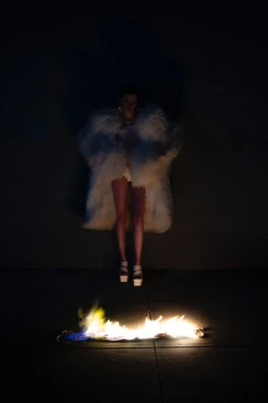 A woman wearing a large, white fur coat and heels stands in front of flames in a fashion or editorial photoshoot in a dark environment.