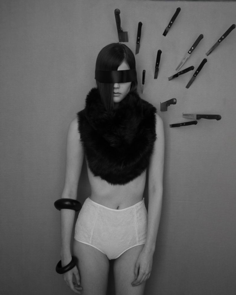 Black-and-white portrait of a woman with a black blindfold and a fur shawl, knives are sticking to the wall behind her.