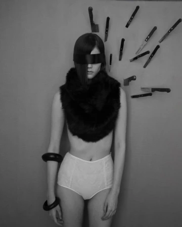 Black-and-white portrait of a woman with a black blindfold and a fur shawl, knives are sticking to the wall behind her.